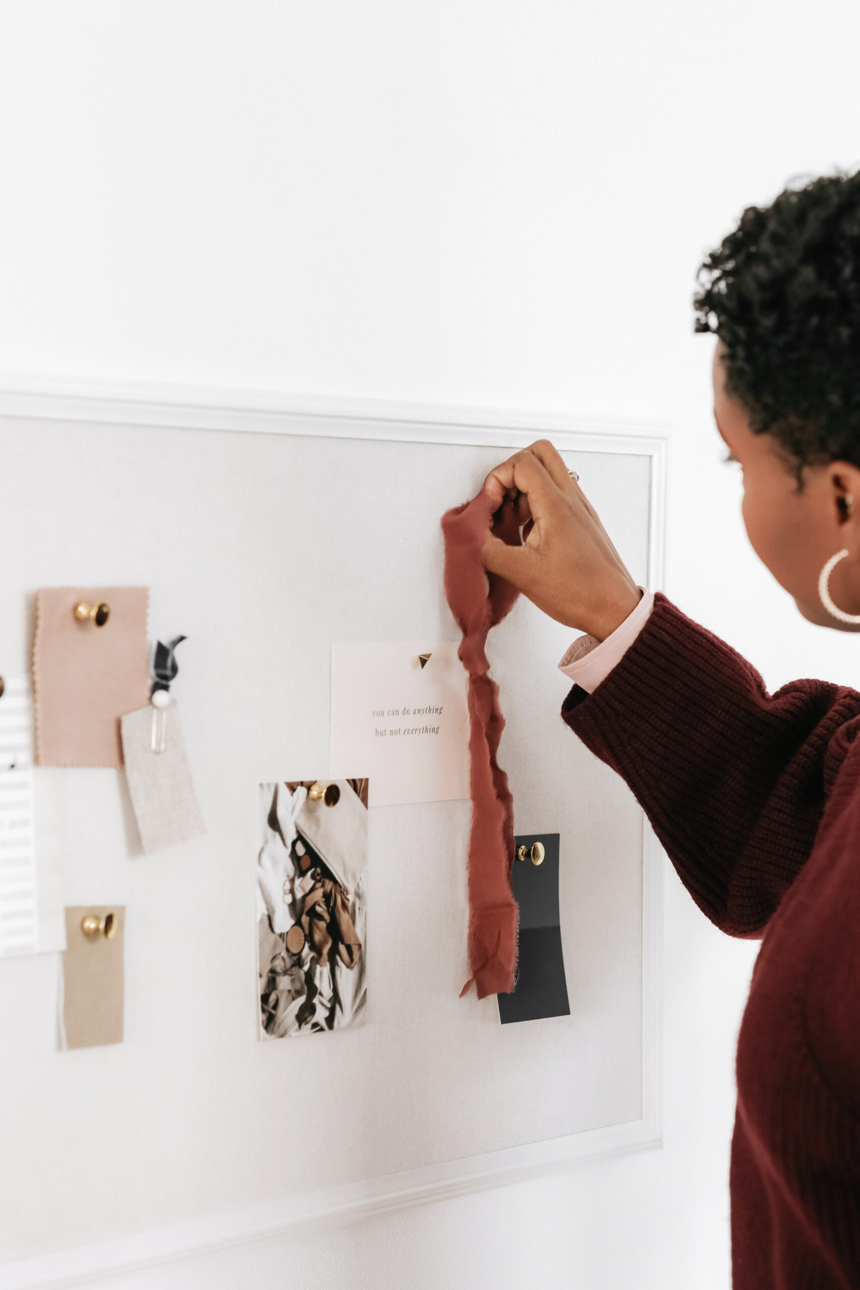 a woman wearing a burgundy knitted jumper and gold earrings building a brand moodboard on a wall, featuring a strip of clay coloured linen, some tonal colour swatches, typography postcards and inspirational imagery